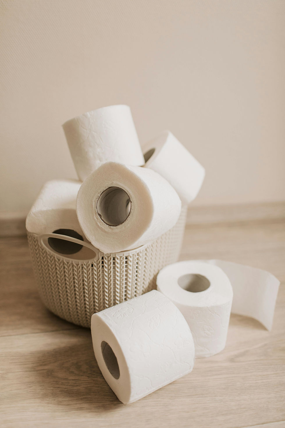 Toilet paper rolls in a beige woven basket on a light wood floor. Neutral-toned background, simple and tidy setting.