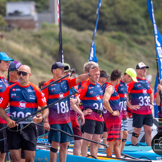 People in red and blue sports jerseys with numbers stand by paddleboards on a beach, some chatting or gesturing, with flags in the background.