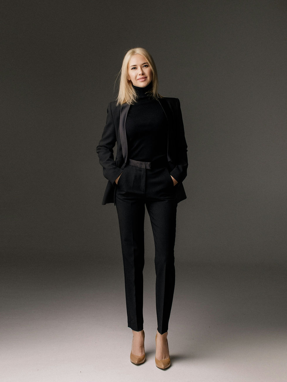 Woman wearing black suit smiling at camera with her hands in pockets.