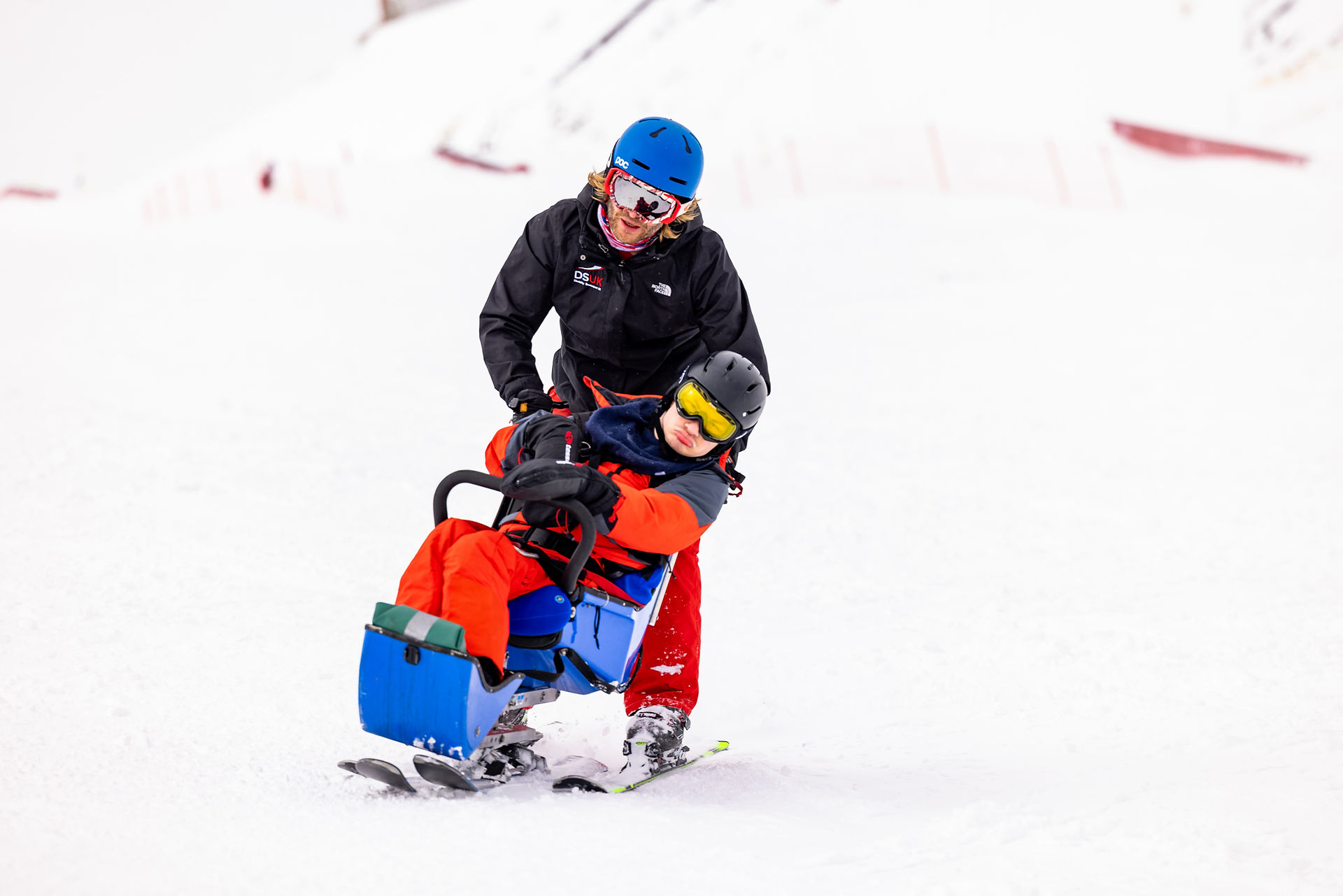 A male ski instructor skiing behind a male sit skier holding the back of his seat as they ski down the mountain together.  