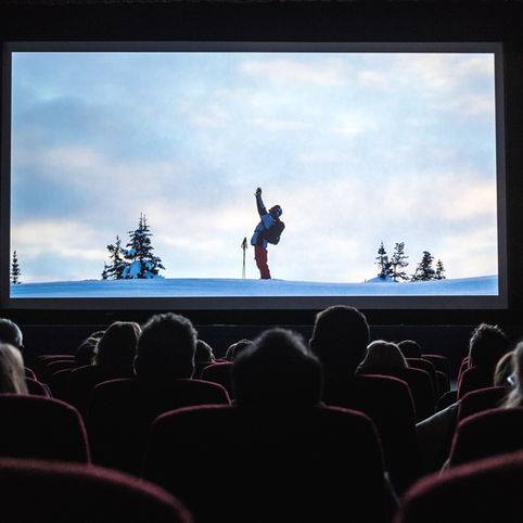Audience watches a ski climber in blue and red on a snowy ridge on a theater screen, framed by dark seats and dim curtains