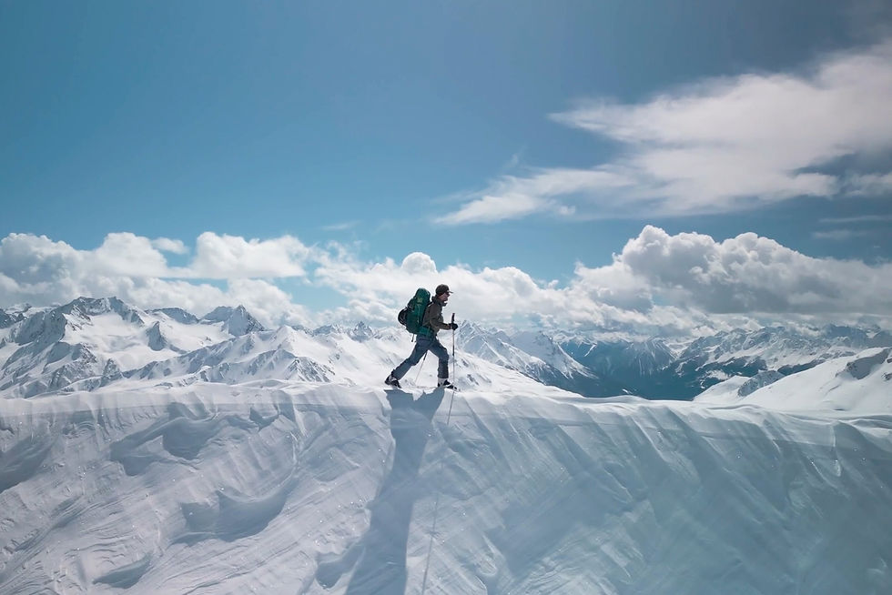 Lone mountaineer with backpack and poles crossing a snowy ridge under a bright blue sky, with vast mountain peaks behind