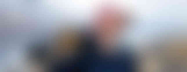 A man with a white beard holding a pair of skis over his shoulder by the side of a road with the ocean in the background