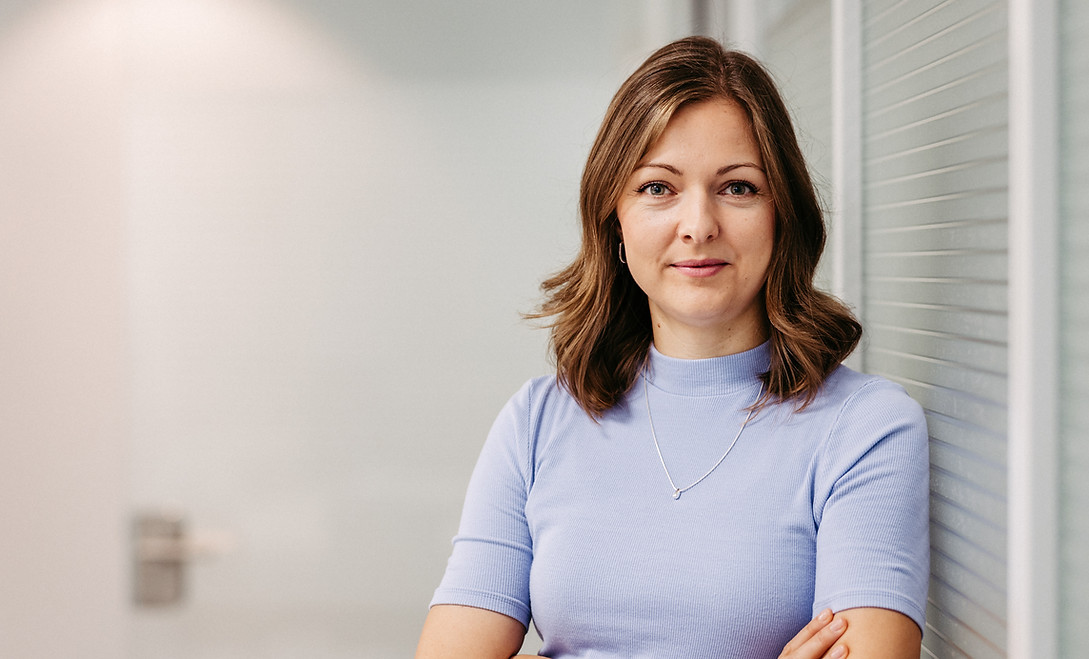 Frau trägt ein lilafarbenes Tshirt und lehnt sich seitlich an eine Wand, Arme verschränkt