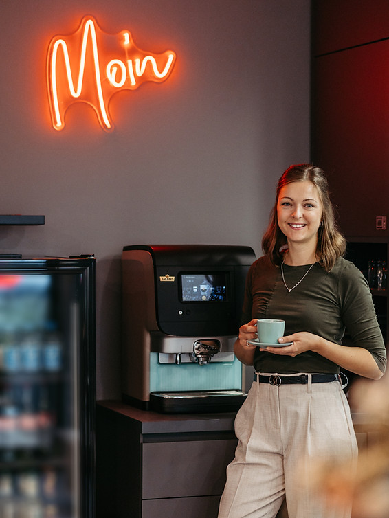 Frau mit Kaffeetasse in der Hand steht neben dem Kaffeeautomaten