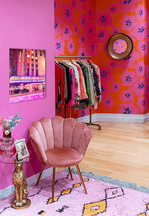 Pink room with colorful floral wallpaper, a clothes rack, and a plush pink chair. A framed Radio City Music Hall art piece and a small side table are visible.