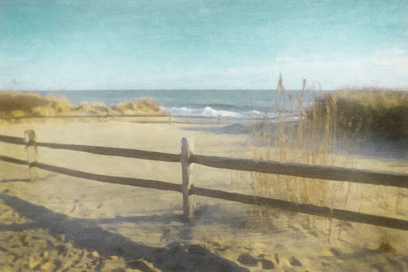 A sandy beach with a wooden fence, dune grasses, and calm blue ocean waves under a clear sky. Mood is serene and peaceful.