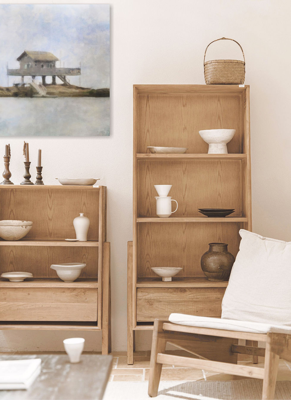Minimalist room with wooden shelves holding pottery and candles. A painting of a stilt house hangs on the wall. Calm, earthy tones.