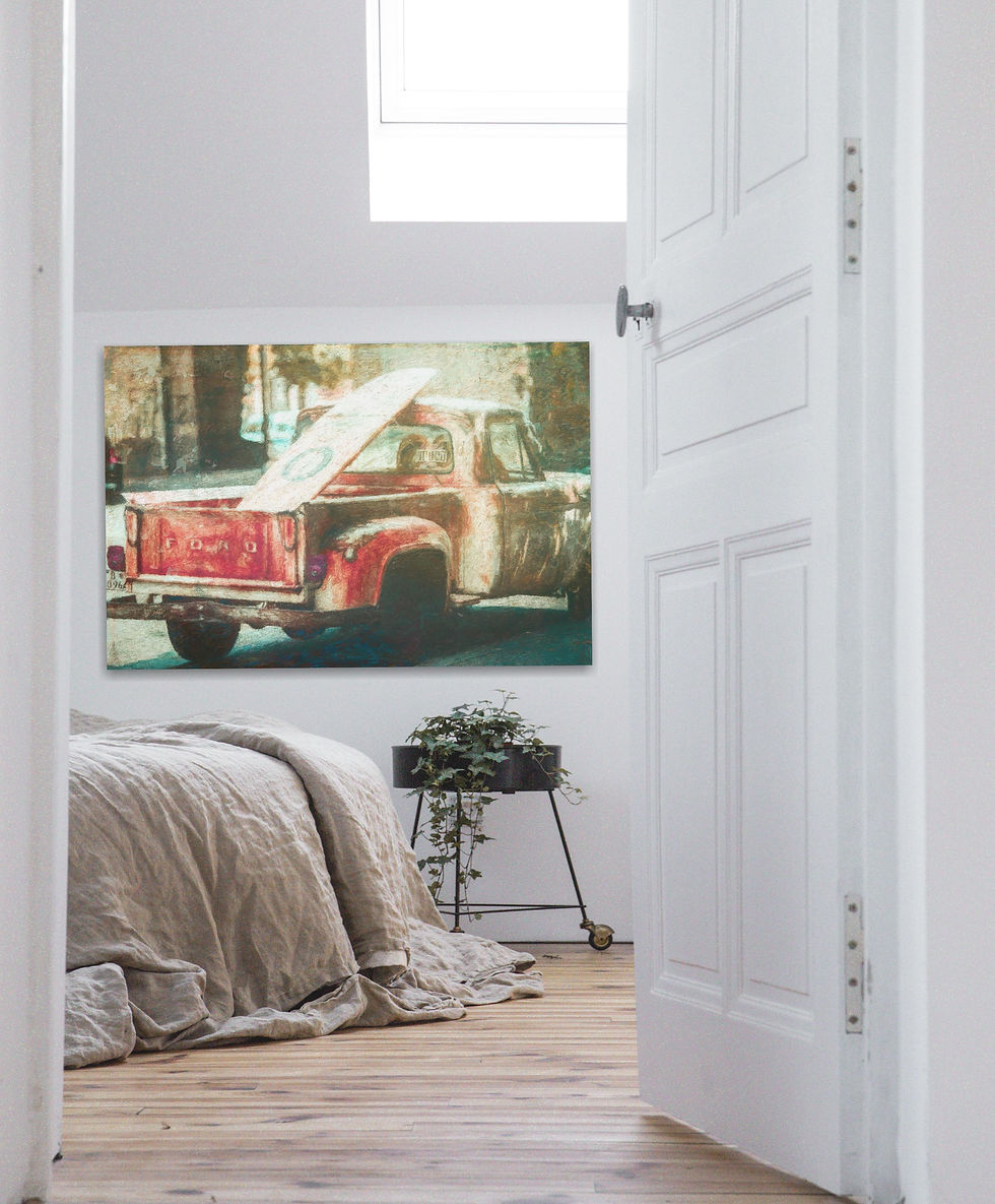 Bedroom with a rustic vibe; a bed with beige linens, plant on a stand, and art of a red truck and surfboard on the wall, under skylight.