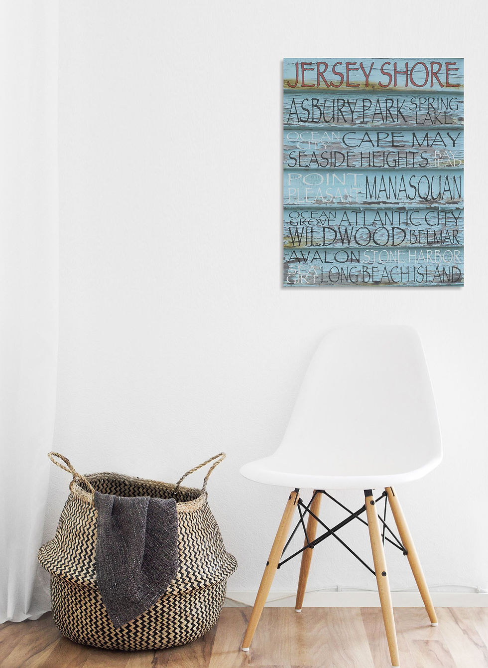 White room with a wicker basket and white chair on hardwood floor. Wall art lists beach names, titled "Jersey Shore," in blue and red.