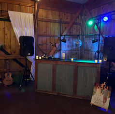 Image shows a DJ booth with a speaker on a stand on either side. Blue and green lights shine down from behind. The booth sits in the corner of a rustic barn  venue.