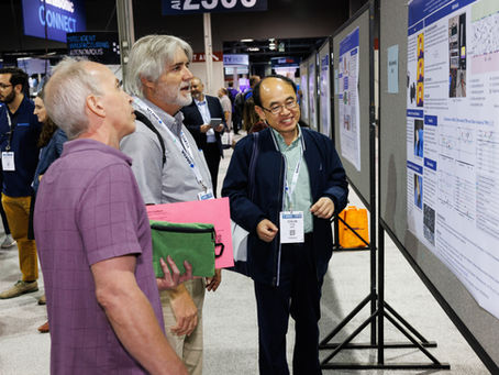Attendees engage in a discussion at a technical conference, examining research posters and exchanging ideas in a bustling exhibit hall.