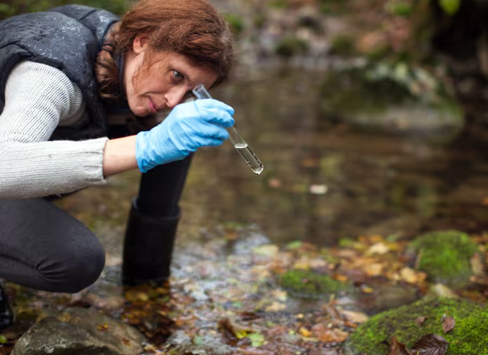 A woman near a river wearing rubber gloves and holding a test tube of water after collecting a sample from the river.