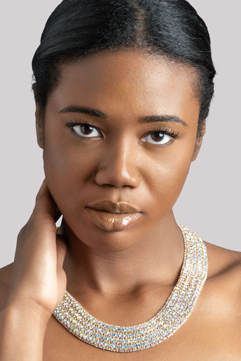 Close-up color portrait of a woman with glossy lips and a serious expression, wearing a sparkling beaded necklace and gently touching her neck.