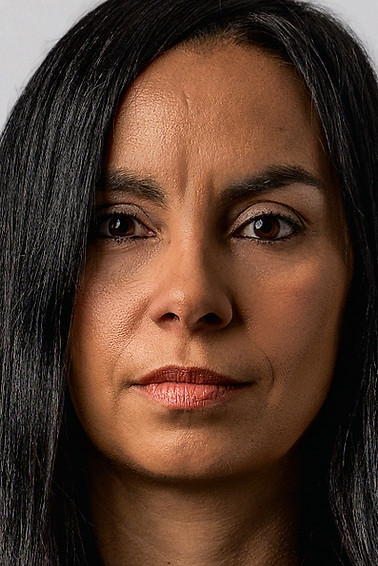 Professional executive headshot of a poised woman with long dark hair wearing a black blazer and neutral blouse against a light gray background, facing forward with a steady, confident expression that communicates authority and credibility.