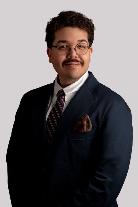 Professional portrait of a man wearing glasses, a dark suit, white shirt, striped tie, and patterned pocket square, posed against a neutral gray background.