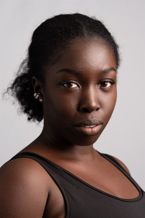 Close-up portrait of a woman with natural hair pulled back, wearing a black tank top, against a light gray background.