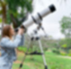 Jovem sorridente observando o céu com um telescópio refrator montado em tripé, em um ambiente ao ar livre com árvores e gramado ao fundo, demonstrando interesse por astronomia durante o dia.