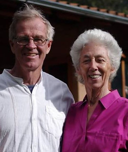 An older Susan with short white hair, smiling wearing a pink button down shirt, and an older man gray hair with glasses