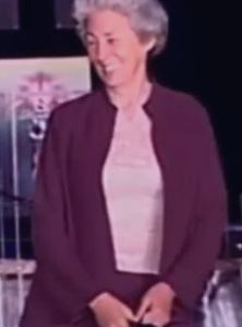 An older Susan with short white hair, smiling, wearing a pink blouse and maroon coat and dress