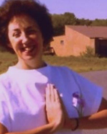 A young Susan in her 20s with short brown hair wearing a white tee shirt holding her hands together in prayer while smiling