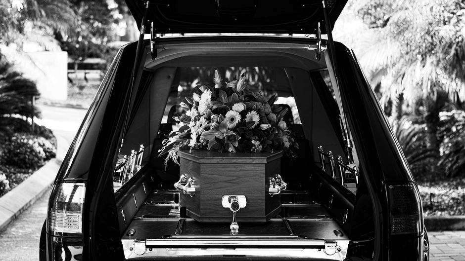 Black and white picture of a hearse’s hatchback is open and we can sea a coffin covered with flowers.