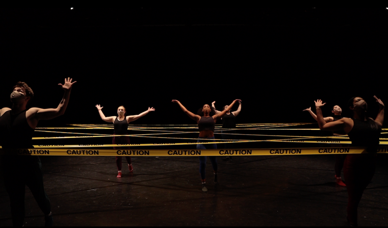 A group of dancers in masks stand with arms spread in a web of caution tape.