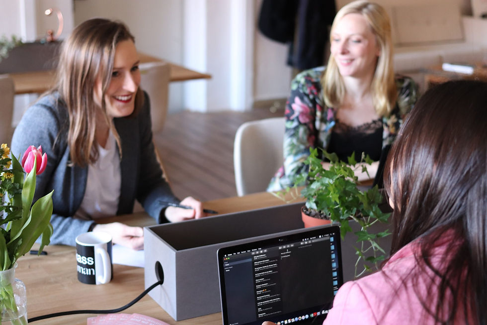Drei junge Frauen sitzen lächelnd an einem Tisch mit Blumenvasen und aufgeklapptem Laptop