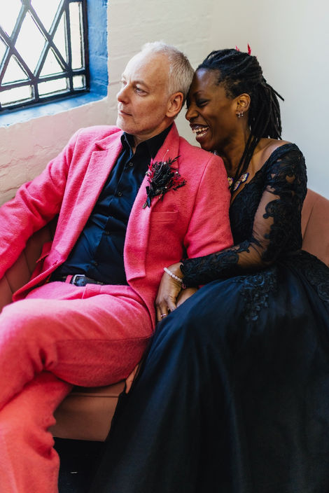 A bride in a black dress sitting with her groom in a pink suit, he has a black dried flower buttonhole