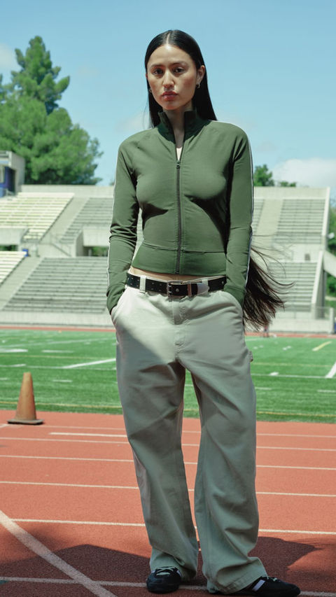 A woman in an olive green zip-up hoodie and khaki pants stands confidently on a running track, blending sporty and casual style.