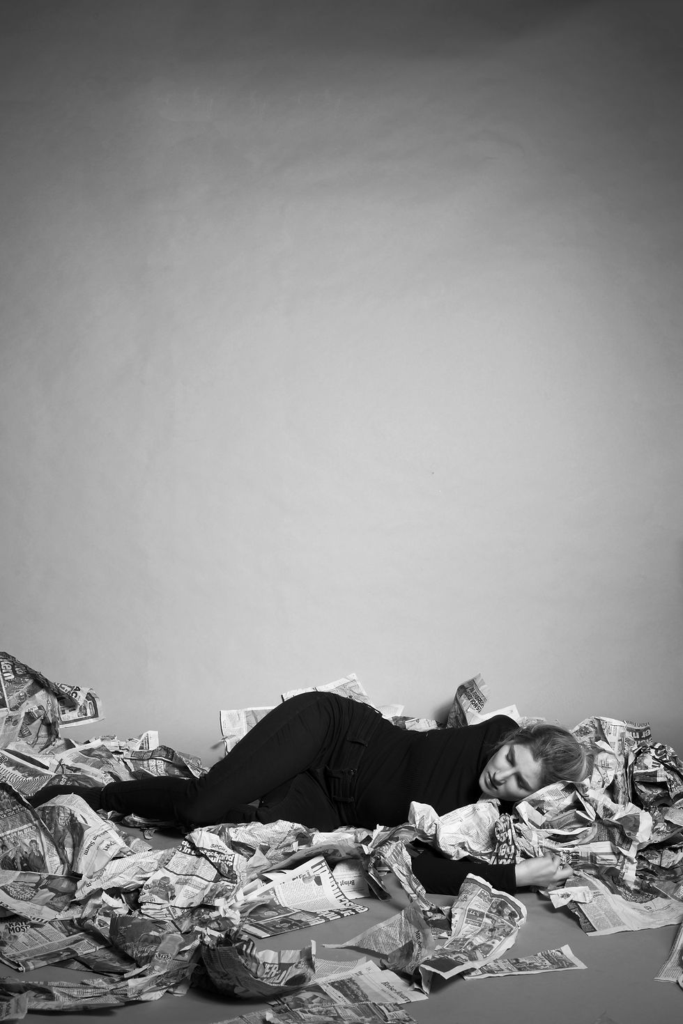 Schwarzweiß Fotografie einer Frau die erschöpft am Boden inmitten von Zeitungen liegt.
