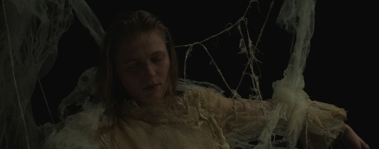 Eine Frau hängt an Spinnweben artigen Fäden mit geschlossenen Augen in einem ansonsten komplett schwarzen Raum.