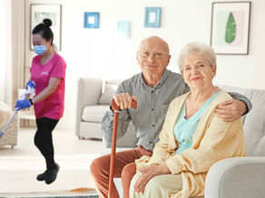 Vanhempi pariskunta istuu sohvalla ja hymyilee kameralle samalla kuin Sivexin siivooja siivoaa lattiaa taustalla.