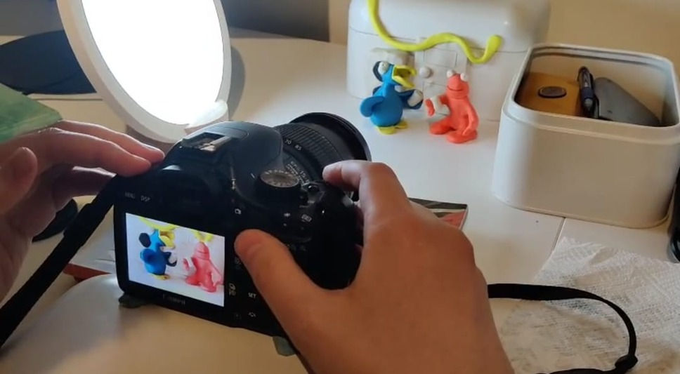 Henkilö säätää kameraa pöydällä kuvatakseen pieniä muovailuvahahahmoja valaistussa sisätilassa.