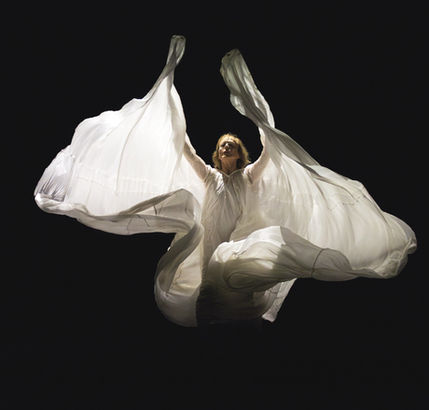 Danseuse en robe blanche ample, bras levés, tissu en mouvement sous une lumière scénique sur fond noir.