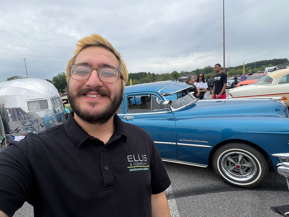 Smiling man in glasses takes a selfie at a car show beside a blue classic car; his black polo reads ELLIS & COMPANY INSURANCE AGENCY