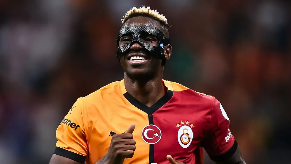 Soccer player in yellow and red jersey with black mask, smiling on field. Galatasaray logo visible. Background is a blurred stadium.