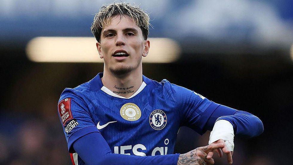 Football player in blue jersey with "Chelsea" logo, adjusting arm bandage. Stadium background, focused expression, FIFA badge visible.
