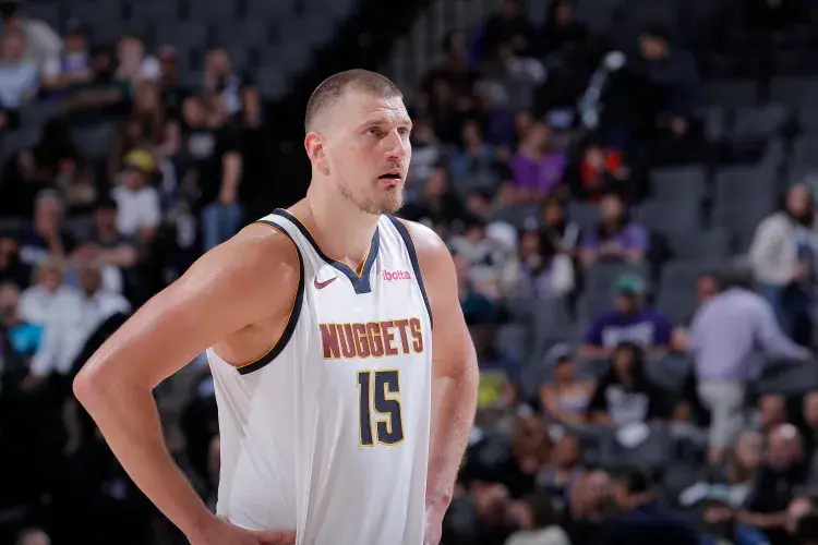 Basketball player in white "Nuggets" jersey number 15 stands focused on court. Blurred audience in background. Action and concentration.