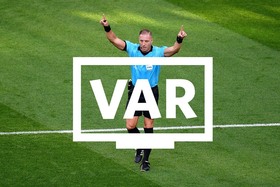 Soccer referee in blue raises both index fingers on a green field as a large VAR sign overlays him.
