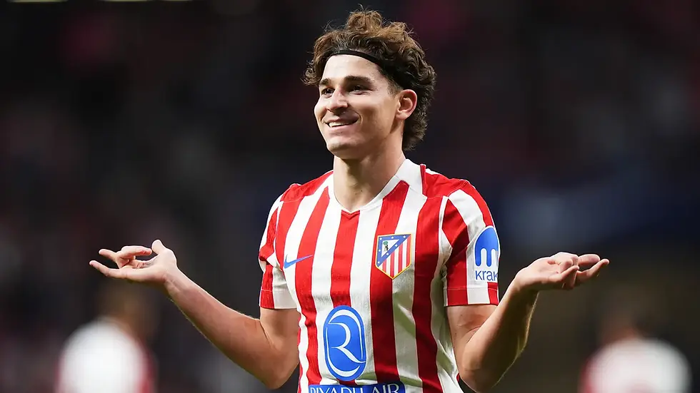Smiling soccer player in red and white striped jersey with arms out, on field. Badge and sponsor logos visible. Dark, blurred background.
