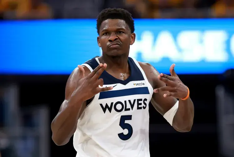 Basketball player in white "Wolves" jersey with number 5 makes gesture on court, blue and orange background. Determined expression.