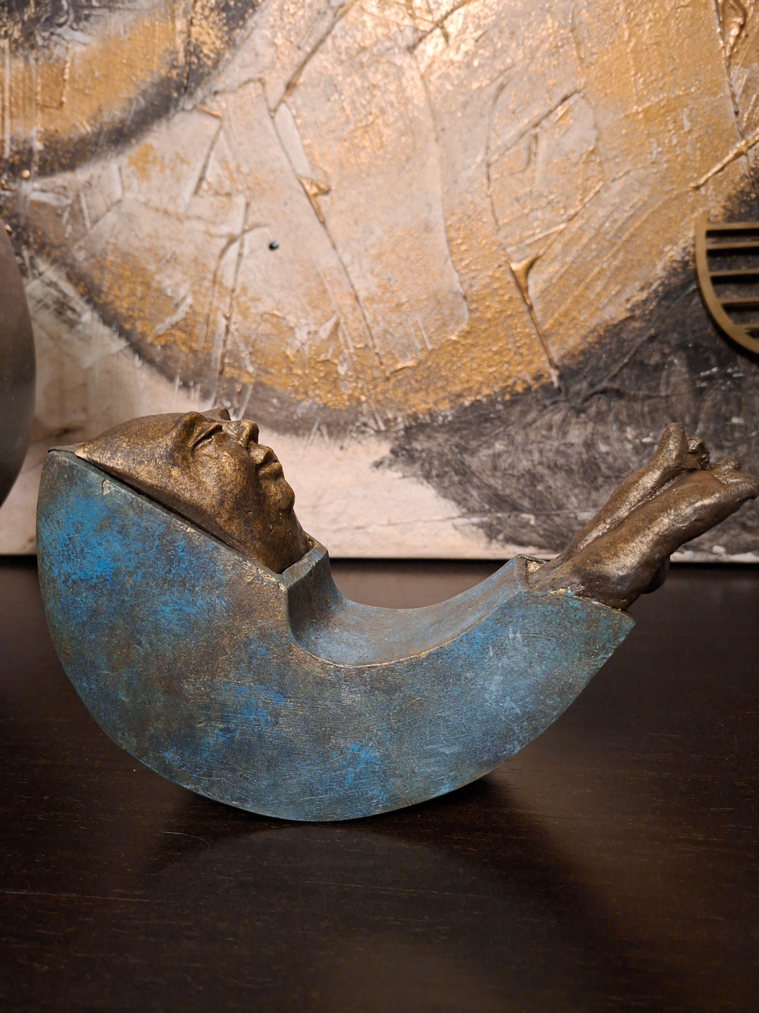 Figura humana de bronze em berço azul, com arte abstrata ao fundo.