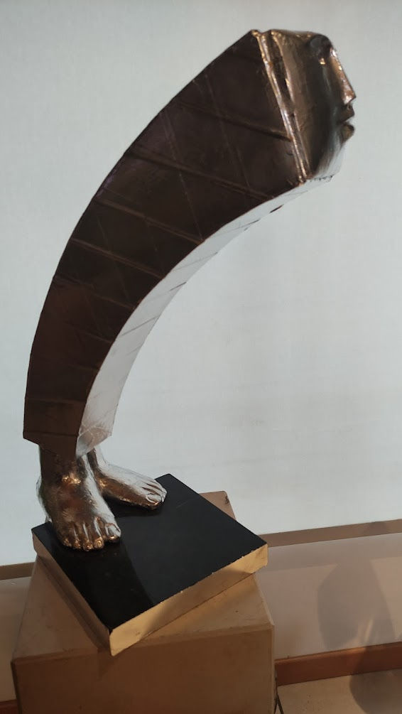 Miniaturbild: Moderna escultura abstrata de bronze, figura humana curvada sobre base preta.