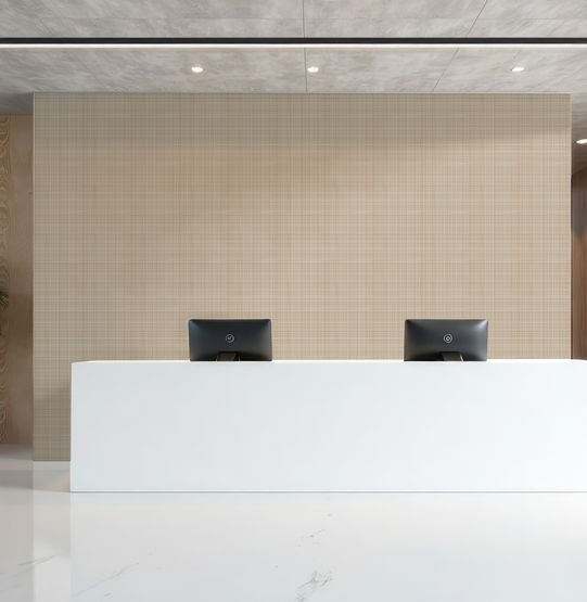 A modern reception area features a long white desk, dual monitors, a potted plant, and wood-panelled walls accented with Covr’s Woven Wall wallpaper in Caramel, all illuminated by bright natural light.