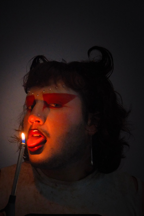 portrait photo of a male model with devil-ish make up holding a fire lighter to his face and tongue
