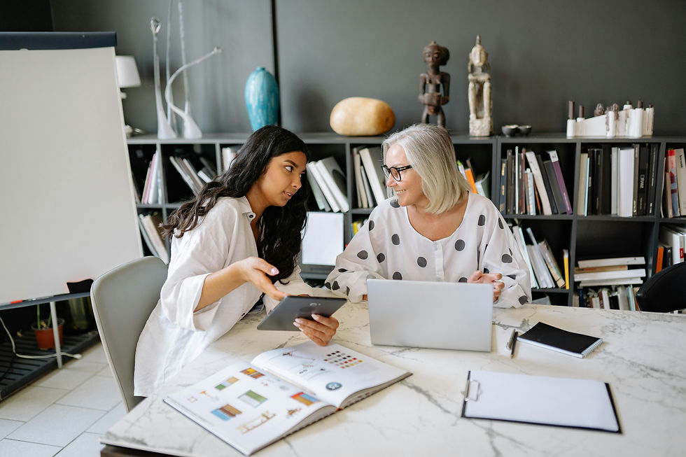 Two professional women, one with long dark hair and the other with short gray hair and glasses, engage in a discussion at a modern office table. The younger woman holds a tablet and gestures as she explains something, while the older woman, working on a laptop, smiles and listens attentively. The office features bookshelves filled with books and decorative sculptures, creating a collaborative and creative atmosphere. This image represents a supportive work environment, where inclusive leadership and open communication foster success for neurodivergent individuals by ensuring clarity, respect, and teamwork.