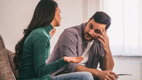 A frustrated man rubs his forehead while a woman talks to him on a couch — illustrating a perseverative thought loop playing out between a parent and their neurodivergent adult child