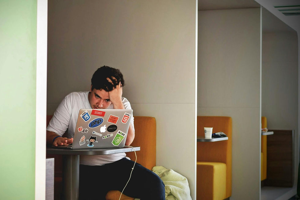 Autistic man in a white shirt stressed, holds head, works on a sticker-covered laptop in a booth. Neutral colors, orange seats, minimal decor.