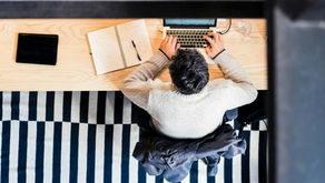 A person wearing a beige sweater sits at a wooden desk, typing on a laptop. An open notebook with handwritten notes and a pen rests beside them, along with a tablet and a glass of water. The overhead perspective emphasizes focus and concentration. This image represents the importance of finding meaningful employment, especially for autistic adults, highlighting how the right job can contribute to financial independence, confidence, and personal fulfillment.